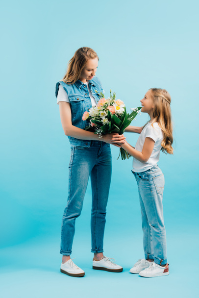 full length view of child in white t-shirt and jeans presenting flowers to mother in denim clothes on blue - Photo, Image