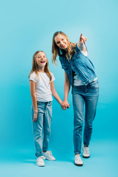 full length view of excited girl holding hands with mother pointing with finger and looking away on blue - Foto, imagen