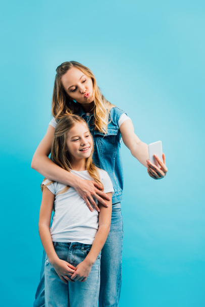 young woman pouting lips while taking selfie on smartphone with daughter wearing white t-shirt and jeans isolated on blue - Foto, imagen