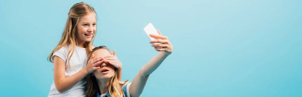 girl covering mothers eyes with hands while she taking selfie on smartphone isolated on blue, panoramic shot - Photo, Image