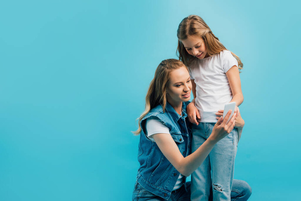 junge Frau in Jeansweste benutzt Smartphone, während sie in der Nähe ihrer Tochter hockt, isoliert auf blauem Grund - Foto, Bild