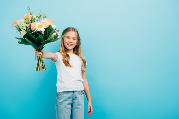 girl in white t-shirt and jeans holding bouquet in outstretched hand while looking at camera isolated on blue - Fotografie, Obrázek