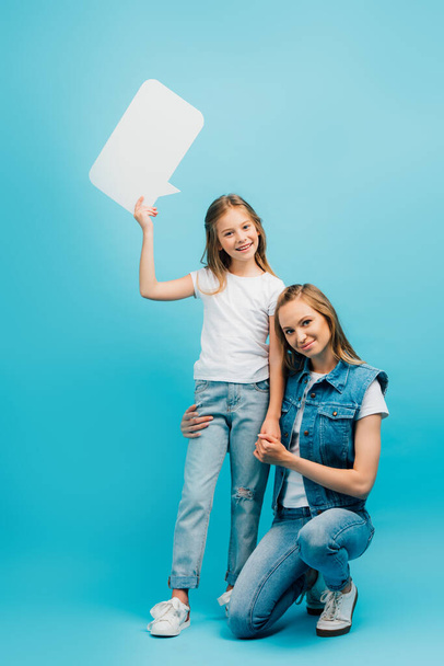 young woman in denim clothes squatting near daughter wearing white t-shirt and jeans holding speech bubble on blue - Foto, immagini