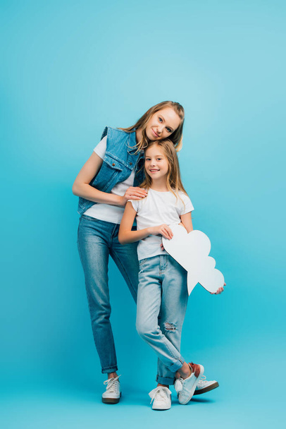 full length view of young woman in denim clothes touching daughter holding thought bubble on blue - Foto, Imagen