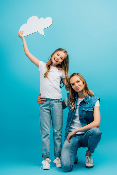 молодая женщина в джинсовой одежде приседает рядом с дочерью в белой футболке и джинсах в джинсах, держа мыльный пузырь на синем - Фото, изображение
