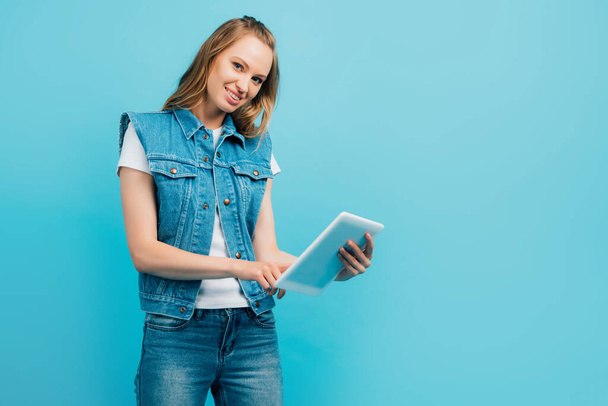 young woman in denim clothes using digital tablet while looking at camera isolated on blue - Foto, Imagem