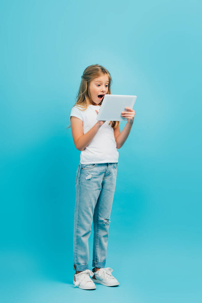 full length view of shocked girl in white t-shirt and jeans using digital tablet on blue - Photo, Image