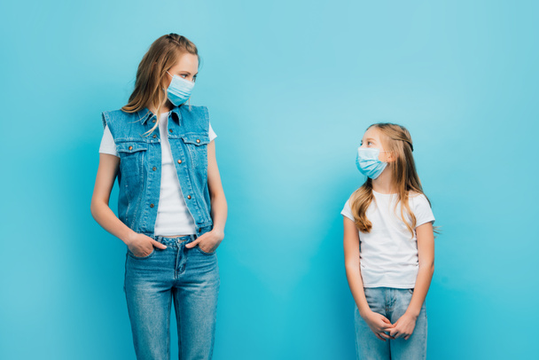 madre e hijo en ropa de mezclilla y máscaras médicas mirándose mientras están aislados en azul - Foto, Imagen