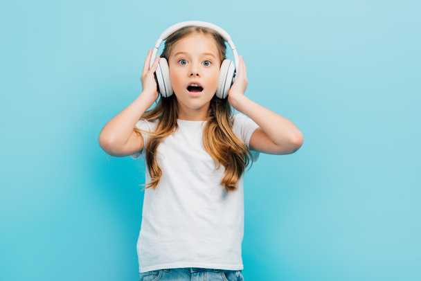 excited girl in white t-shirt touching wireless headphones while looking at camera isolated on blue - 写真・画像