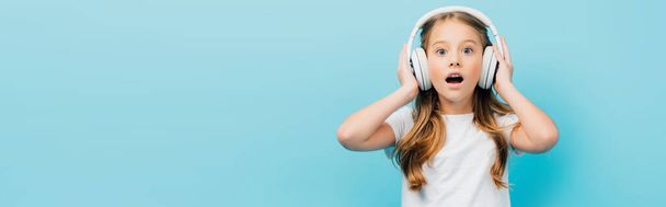 σοκαρισμένη κοπέλα σε λευκό t-shirt αγγίζοντας ασύρματα ακουστικά που απομονώνονται σε μπλε, πανοραμική έννοια - Φωτογραφία, εικόνα