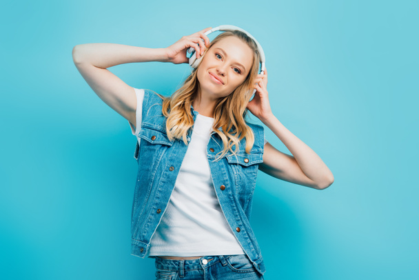 young woman in denim west looking at camera while touching wireless headphones isolated on blue - Zdjęcie, obraz