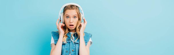 panoramic concept of shocked woman in denim vest touching wireless headphones while looking at camera isolated on blue - Foto, Imagen