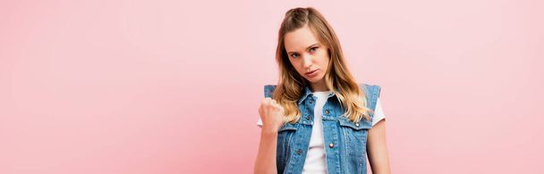 horizontal image of angry woman in denim vest showing clenched fist at camera  isolated on pink - Zdjęcie, obraz