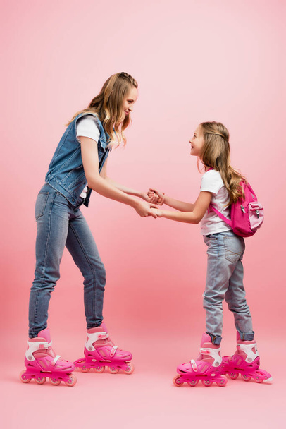 mother and daughter in denim clothes holding hands and looking at each other while standing on roller skates together on pink - Фото, изображение