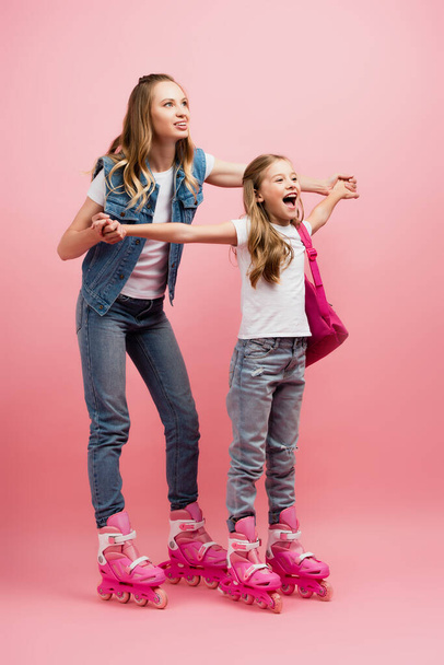full length view of mother holding hands of excited daughter while standing on roller skates together on pink - Foto, Imagem