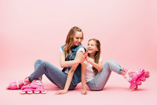 mother and daughter in denim clothes sitting on floor in roller skates and giving high five on pink - Fotoğraf, Görsel