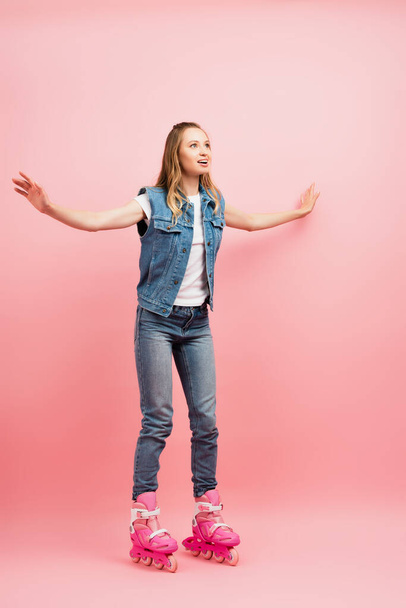 full length view of excited woman in denim clothes and rollers skates with outstretched hands on pink - Photo, Image