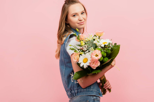 young woman in denim clothes holding bouquet and looking at camera isolated on pink - Foto, Imagen
