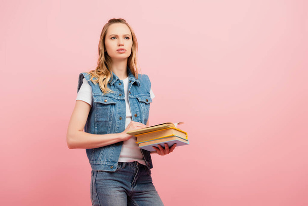 jeune femme réfléchie en gilet en denim regardant loin tout en tenant des livres isolés sur rose - Photo, image