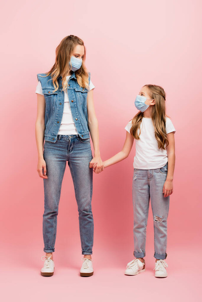 vista completa de la madre y la hija en jeans y máscaras médicas cogidas de la mano y mirándose mutuamente en rosa - Foto, imagen