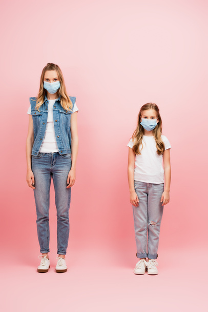 full length view of mother and daughter in medical masks, wearing denim clothes, looking at camera on pink - Foto, Imagen