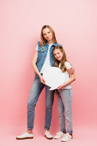 Ganzkörperansicht einer jungen Frau in Jeansweste und Jeans, die ihre Tochter umarmt, während sie eine Gedankenblase auf rosa hält - Foto, Bild