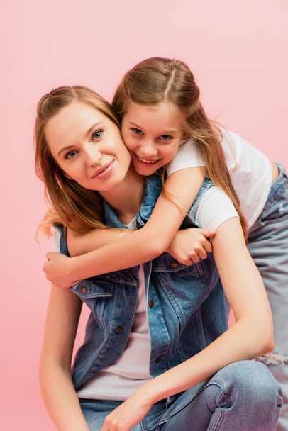 Mädchen umarmt Mutter von hinten, während sie isoliert auf rosa Kamera schaut - Foto, Bild