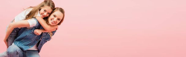 horizontales Bild einer jungen Frau in Jeansweste huckepack Tochter isoliert auf rosa - Foto, Bild