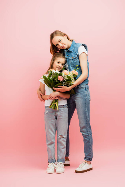 full length view of young woman in denim clothes gifting flowers to daughter and hugging her on pink - Photo, Image