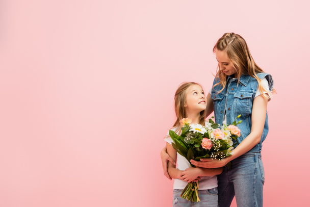 jovem mulher em roupas jeans apresentando buquê de flores para filha isolada em rosa - Foto, Imagem