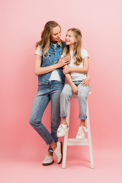 young woman in denim clothes hugging daughter sitting on high stool on pink - Fotó, kép