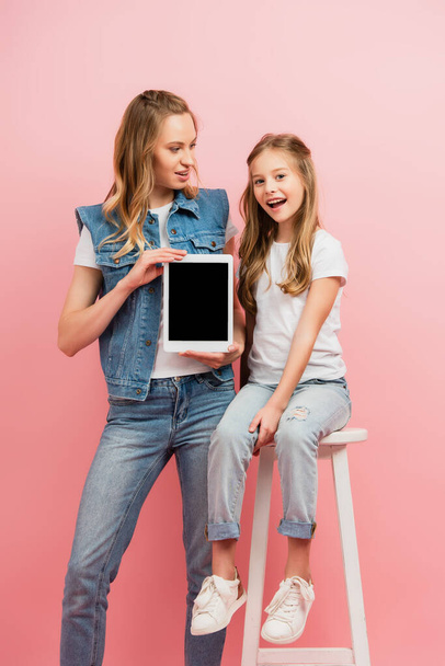 jeune femme en vêtements en denim tenant tablette numérique avec écran blanc près fille excitée assise sur un tabouret isolé sur rose - Photo, image