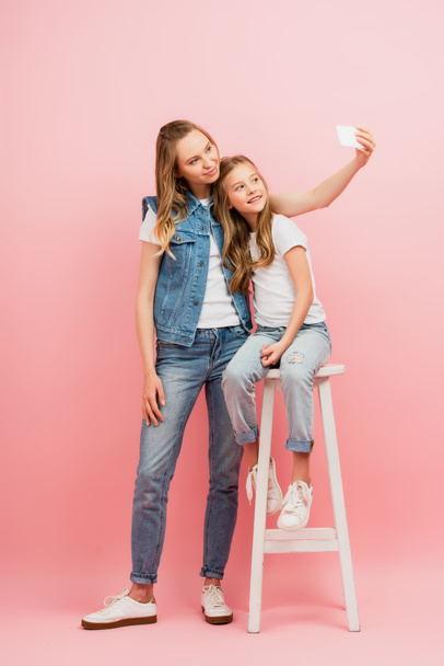 full length view of young woman in denim clothes taking selfie on smartphone with daughter sitting on high stool on pink - Fotó, kép