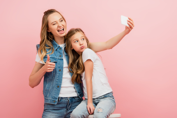 femme excitée en vêtements en denim prendre selfie sur smartphone avec fille boudant lèvres et montrant pouce vers le haut isolé sur rose - Photo, image