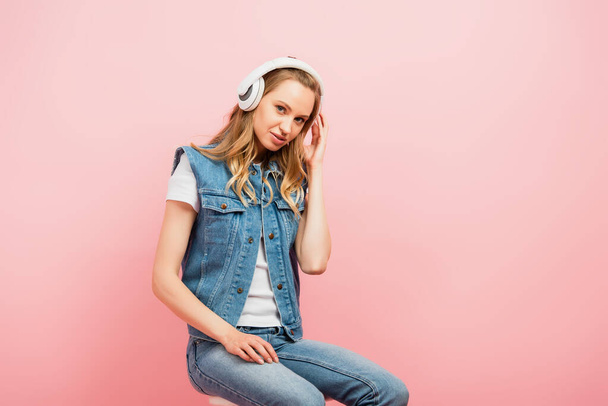 young woman in denim clothes touching wireless headphones while sitting isolated on pink and looking at camera - Foto, Imagen