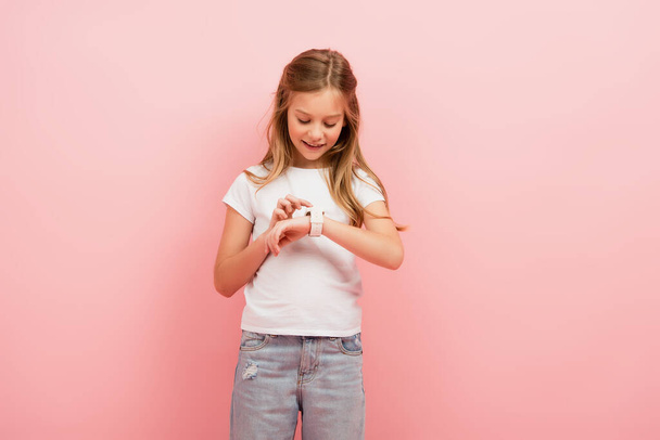 ребенок в белой футболке и джинсах касающихся умных часов, изолированных на розовый - Фото, изображение