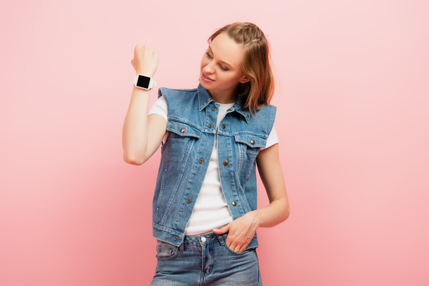 młoda kobieta w dżinsowej kamizelce pokazuje smartwatch na nadgarstku stojąc z ręką w kieszeni odizolowany na różowo - Zdjęcie, obraz