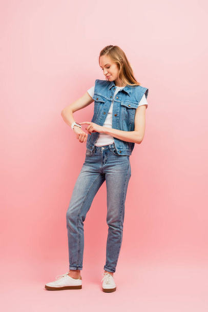 full length view of young woman in denim clothes touching smartwatch on pink - Foto, Imagen
