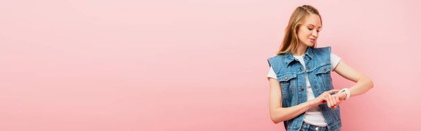 plano panorámico de mujer joven en ropa de mezclilla tocando smartwatch aislado en rosa - Foto, imagen