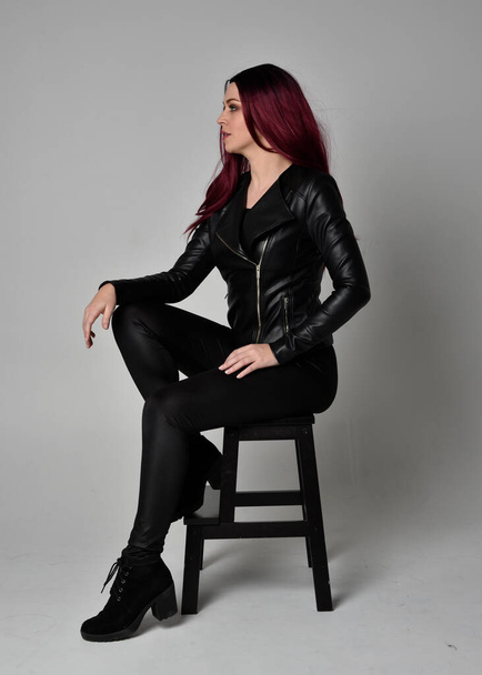 Teljes hosszúságú portré vörös hajú lányról fekete bőrdzsekiben, nadrágban és csizmában. Ülő póz egy széken, elszigetelt egy szürke stúdió háttér. - Fotó, kép