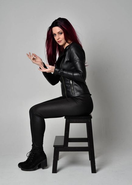 Retrato completo de niña de pelo rojo con chaqueta de cuero negro, pantalones y botas. Posar sentado en una silla, aislado sobre un fondo gris estudio. - Foto, imagen