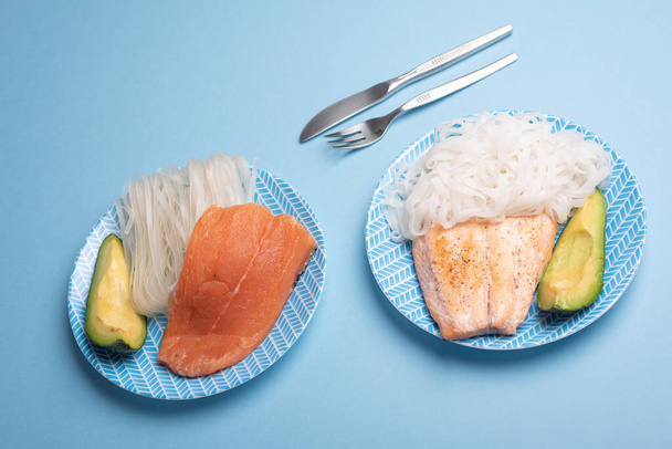 Surowe danie atlantyckiego łososia, makaron ryżowy i plasterek awokado przeciwko ugotowane jeden z nożem i widelcem na zwykłym tle koloru niebieskiego. Gotowy do dodania tekstu - Zdjęcie, obraz