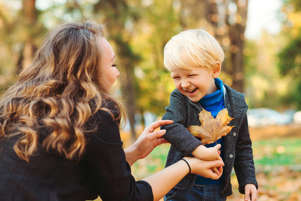 秋の公園で一緒に遊んでいる母親と子供。秋の散歩で幸せな若い家族。秋を楽しむ母と息子。幸せな母親。秋のファッション。落ち葉で楽しむスタイリッシュな家族. - 写真・画像