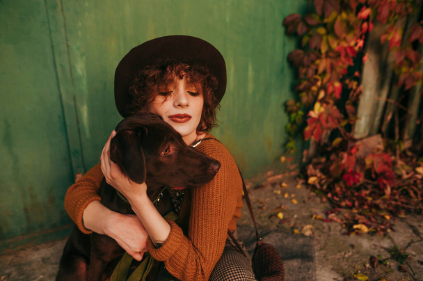 Lähikuva muotokuva kaunis tyttö hattu ja kihara tukka halaus koira istuu syksyllä kentällä kadulla maalaistalon. Lady halaus koira syksyllä maalaismainen tausta. - Valokuva, kuva