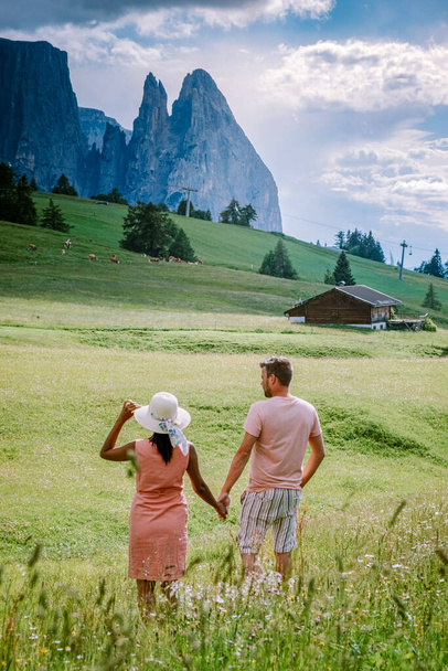 Paar im Urlaub in den Dolomiten Italien, Seiser Alm mit Langkofel-Langkofelgruppe im Hintergrund bei Sonnenuntergang. Gelbe Frühlingsblumen und Holzchalets in - Foto, Bild