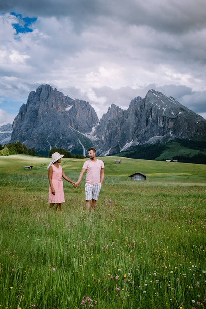para mężczyzn i kobiet na wakacjach w Dolomitach Włochy, Alpe di Siusi - Seiser Alm z grupą górską Sassolungo - Langkofel w tle o zachodzie słońca. Żółte wiosenne kwiaty i drewniane domki w - Zdjęcie, obraz