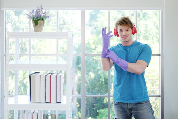 Hymyilevä nuori valkoihoinen mies yllään violetti kumi käsineet puhdas kotitöitä ja rentouttava kuunnella musiikkia punainen kuuloke. Elämäntapa Familyman Loma Concept. - Valokuva, kuva