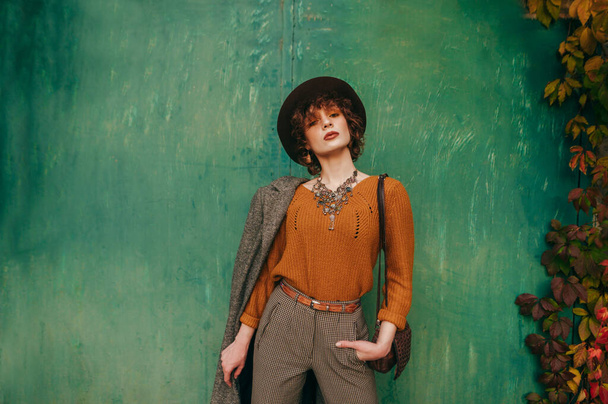 Μόδα πορτρέτο του μοντέλου σε vintage ρούχα σε πράσινο φόντο grunge τοίχο, κοιτάζοντας στην κάμερα με σοβαρό πρόσωπο. Ελκυστική κυρία με μοντέρνα ρούχα που ποζάρει σε έγχρωμο φόντο. Στυλ Vogue - Φωτογραφία, εικόνα