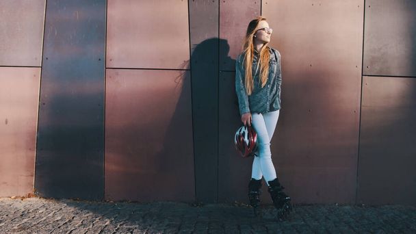 Νεαρή γυναίκα της μόδας καυκάσιος με inline πατίνια και κράνος ασφαλείας ακουμπά στον τοίχο του κτιρίου. Αστική ζωή - Φωτογραφία, εικόνα