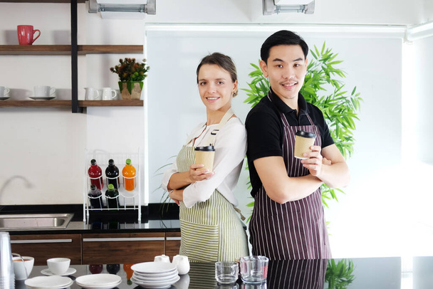 Uśmiechnięta młoda baristka i kelnerzy para kocha Azjatę i białą kobietę trzymających kubki gorącej kawy razem. Uruchomienie kawiarni i koncepcji biznesu kawiarni. - Zdjęcie, obraz